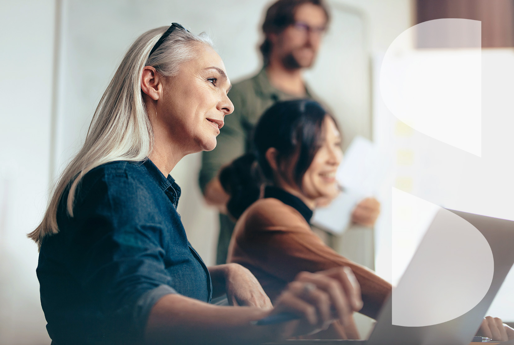 Senior woman with glasses holding her hair back listens to colleagues sitting by during business presentation. Business people meeting in a conference room for a strategy planning.