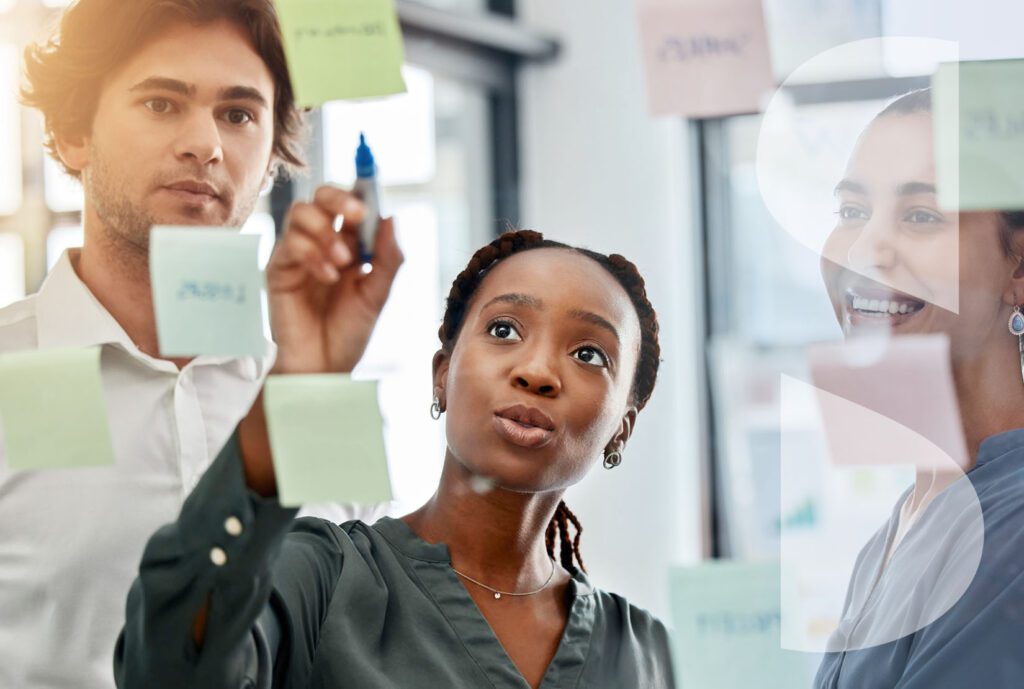 A woman leads a strategy session for a team of 3 using post its on a window.